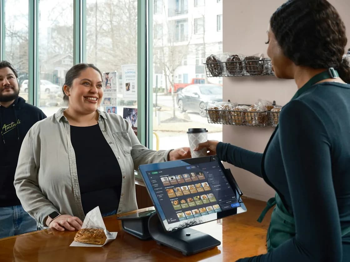 A smiling guest paying at a café counter, a tablet on a stand showing the menu beside a pastry